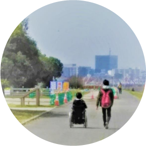 遠くにビル群が見える公園の遊歩道を、車椅子ユーザーと介助者が横並びで進んでいく後姿